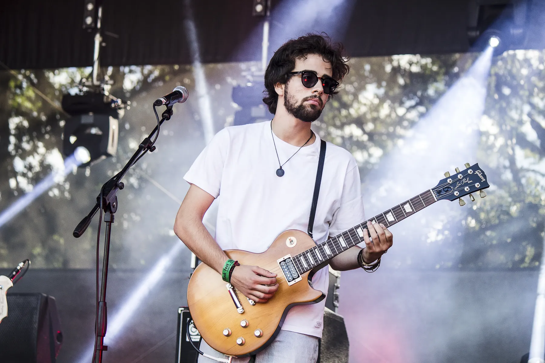 Guitarrista de óculos escuros e camisa branca em palco, entre feixes de luz